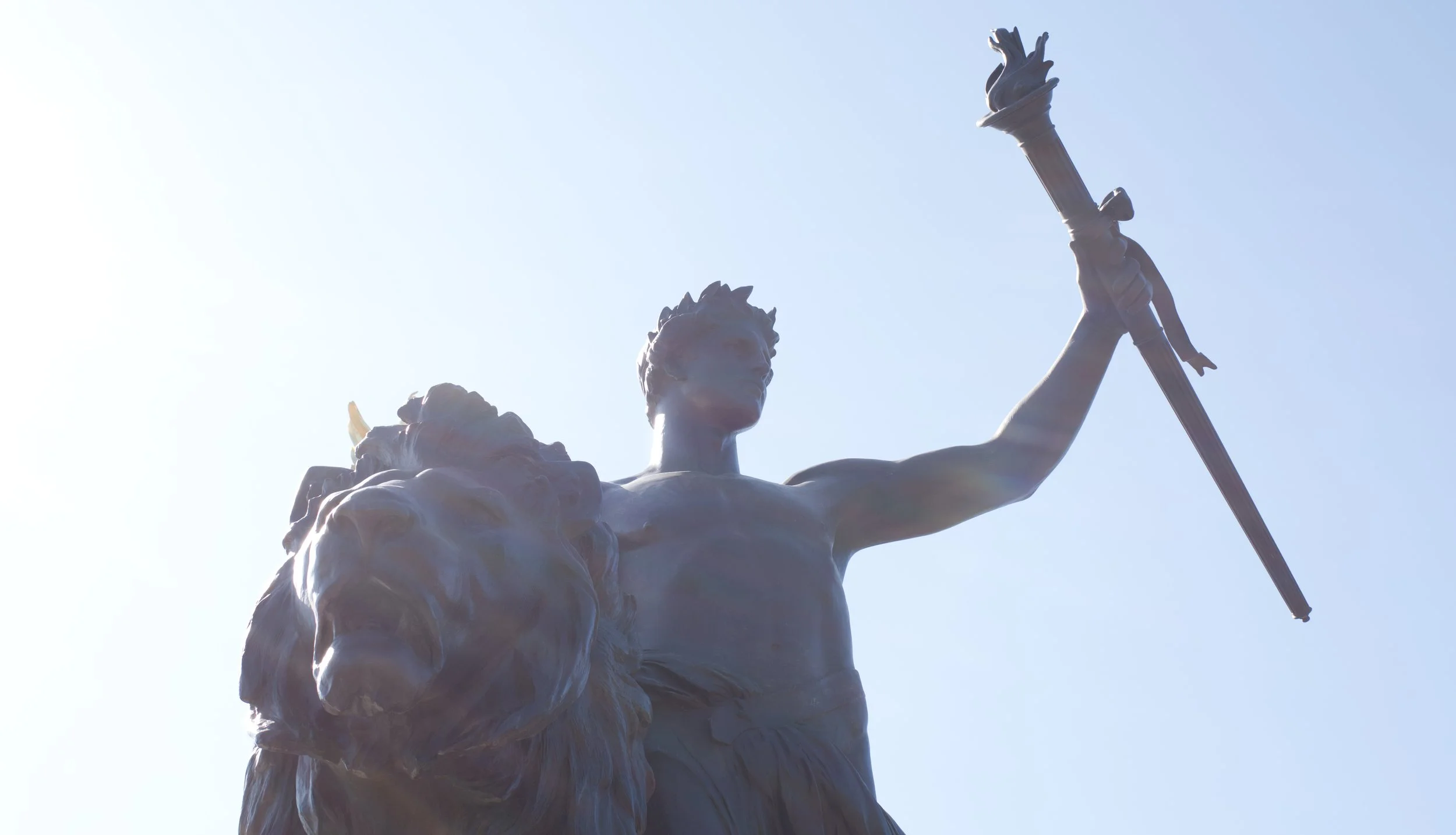 A statue of a man holding a spear with a lion's head on his shoulder, set against a bright sky.
