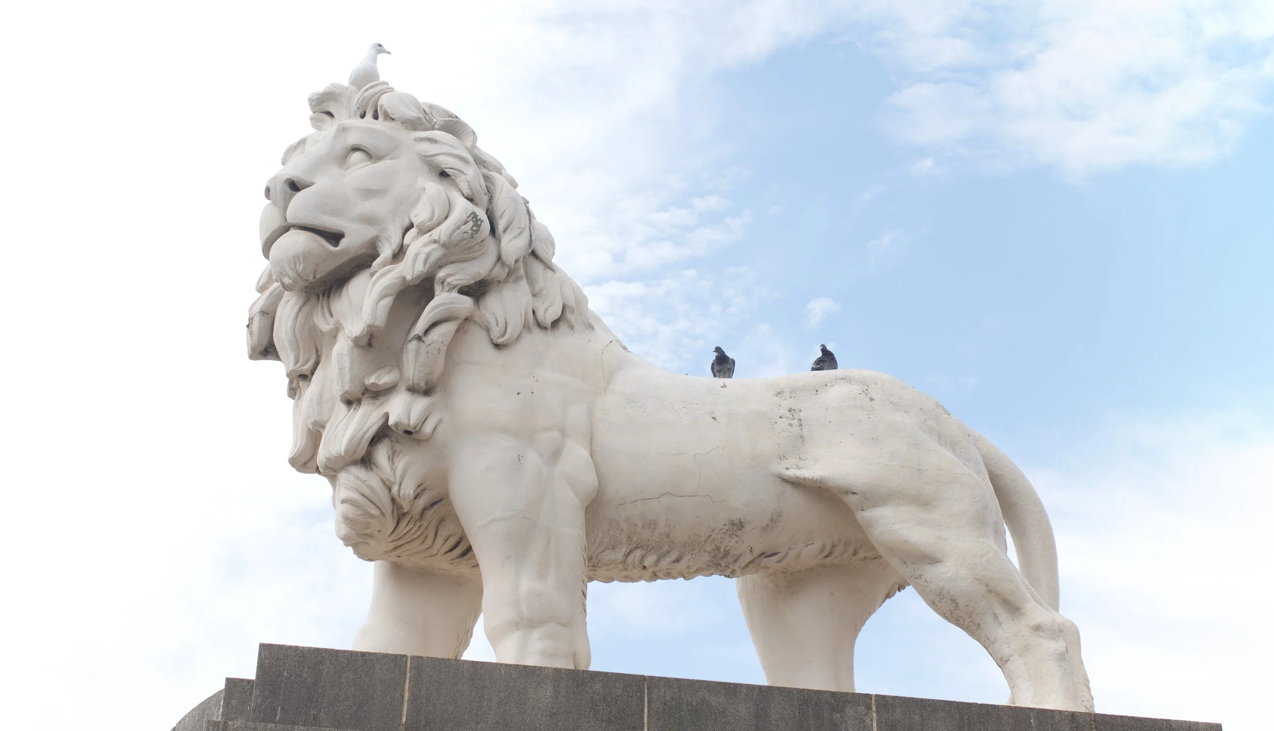 White stone sculpture of a lion with two pigeons resting on its back, set against a partly cloudy sky.