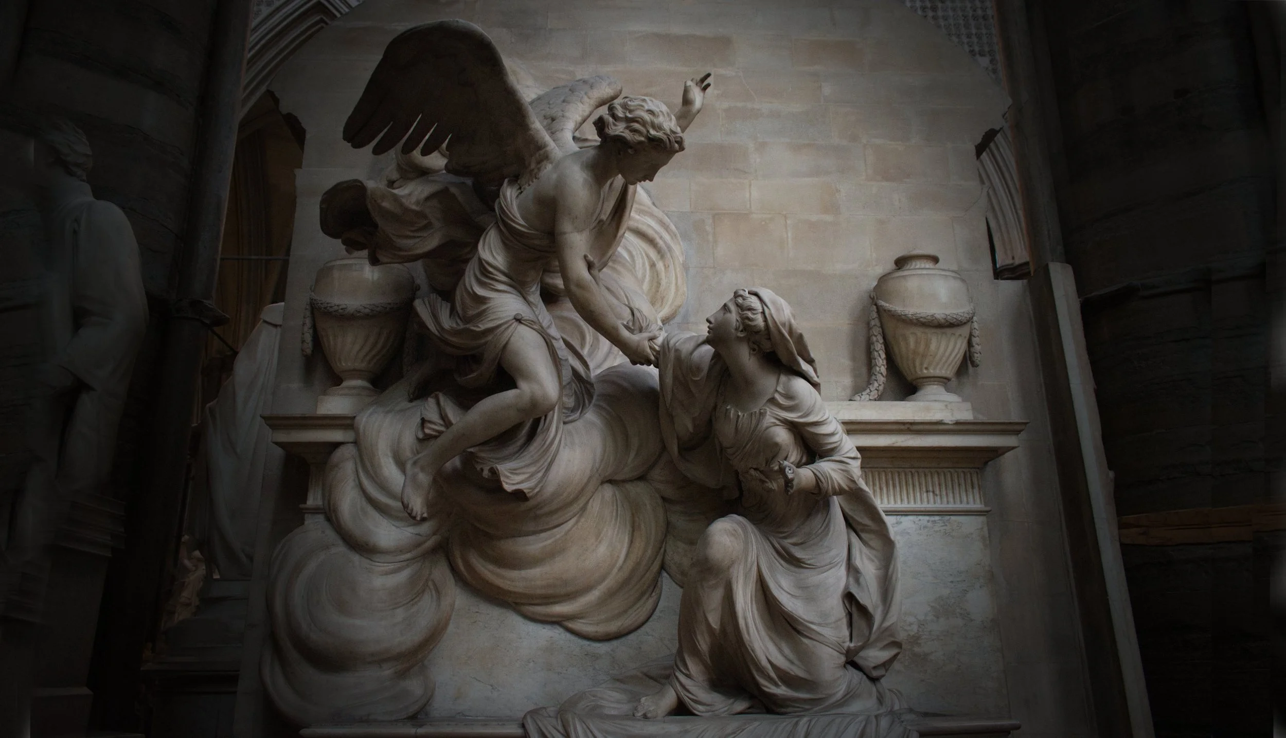 A detailed marble sculpture depicting a dramatic scene with multiple figures, including a winged figure appearing to rescue or support another figure, set against a decorated interior background.