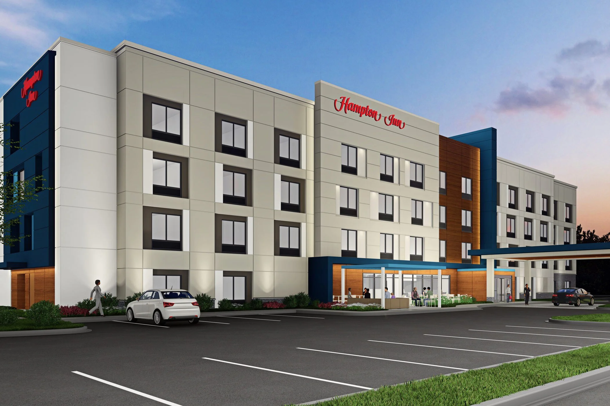 A modern four-story hotel building with the sign 'Hampton Inn' on it, featuring a parking lot in front with several cars and people walking near the entrance, under a blue sky.