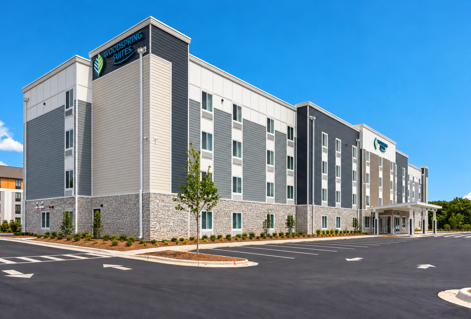 Modern four-story building with a sign that reads 'Woodspring Suites,' featuring gray, white, and beige siding, a stone base, and a parking lot with marked spaces and a few young trees.