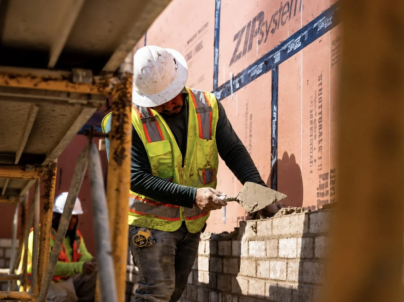 Construction worker in a safety vest and hard hat troweling mortar onto bricks at a building site.