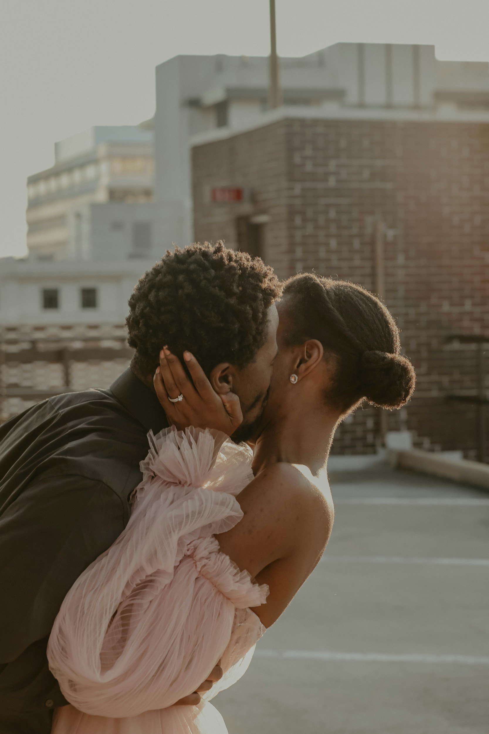 A couple sharing an intimate moment, leaning in for a kiss on a rooftop with city buildings in the background during golden hour.