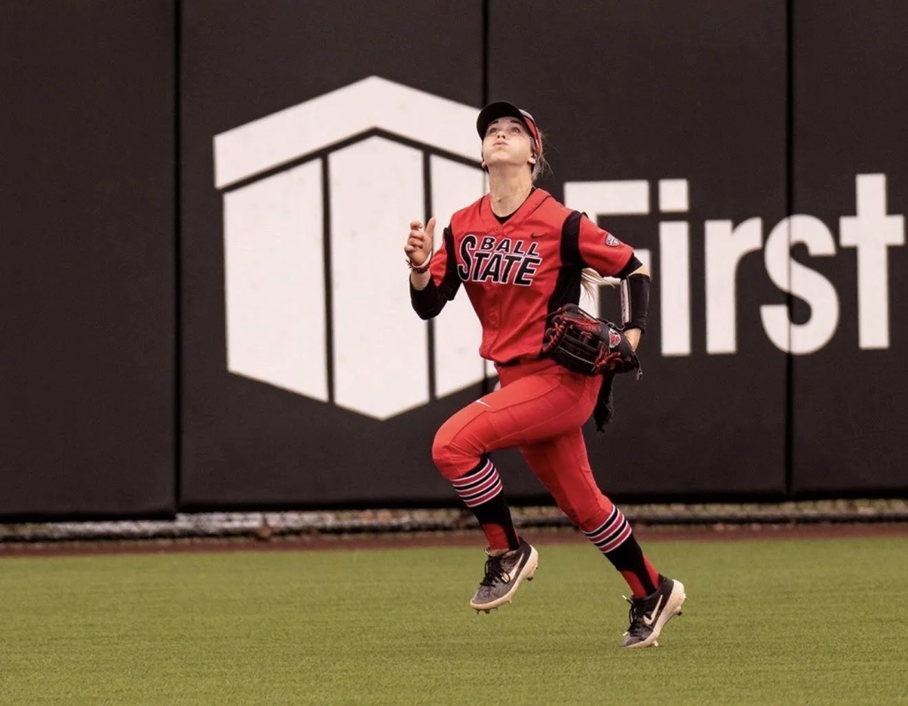 A female baseball player in a red uniform with black accents and the words 'Ball State' on her jersey, running on a baseball field with a black wall and large white logo and text in the background.