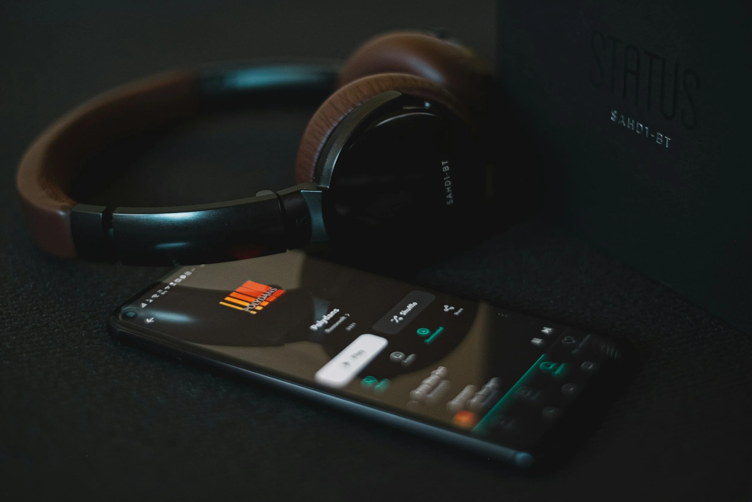 Trådlösa hörlurar av märket Sennheiser, modell HD 1 BT, placeras ovanpå en smartphone med en musikapp öppen, på en svart yta.