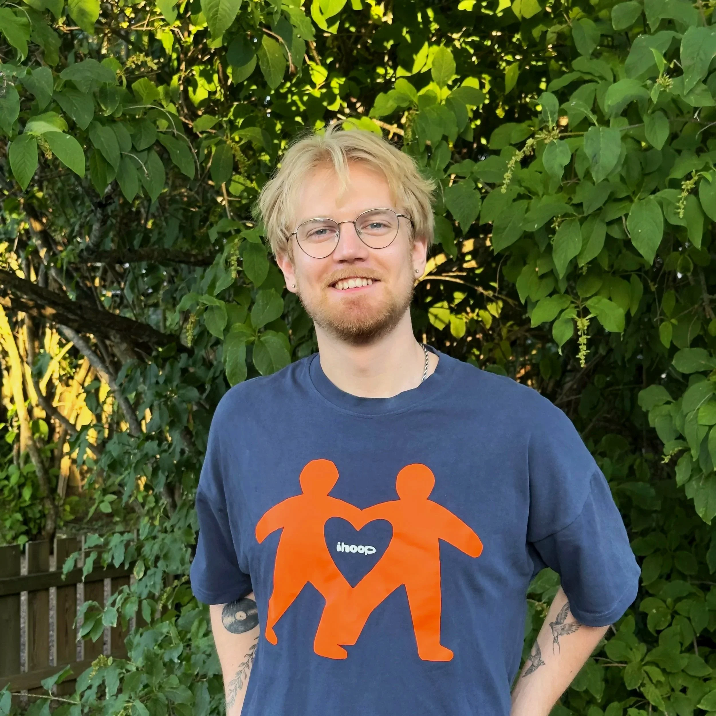 En ung man med blond hår, glasögon och tatueringar på armen, står framför en grön växtvägg och bär en mörkblå T-shirt med en orange logo från Ihoop.