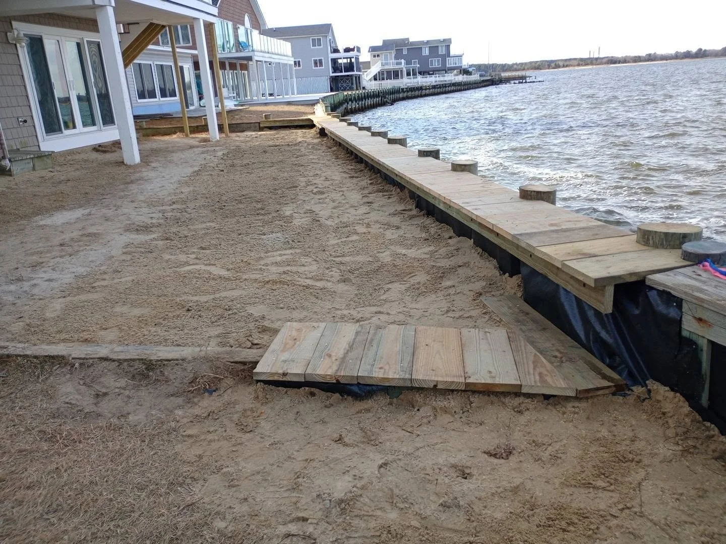 Lakeside construction site with a wooden dock being built along the shore, with sand and sandpaper in the foreground, residential buildings in the background, and water on the right side.