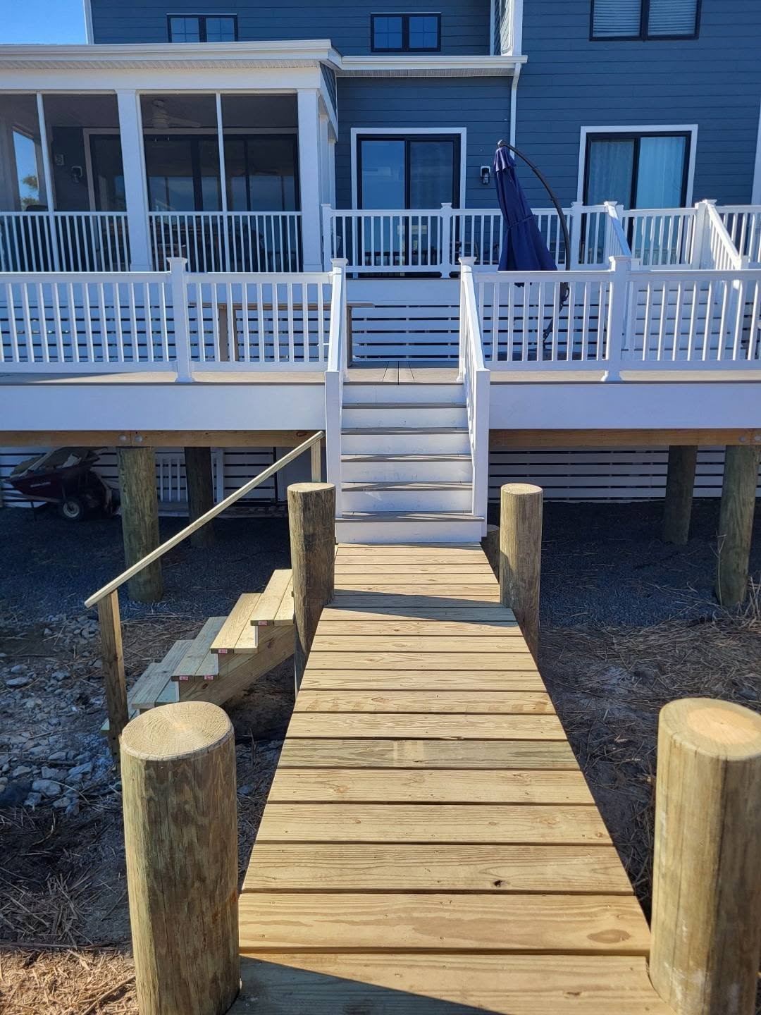 View of a wooden dock leading to a raised blue house with a porch, white railing, and sliding glass doors.