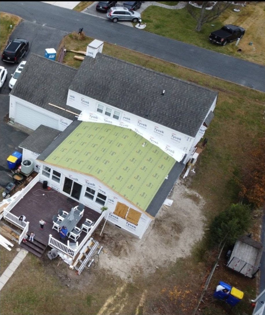 A house under construction with part of the roof covered with yellow insulation material and the other part with dark shingles. The front porch is finished with white railing and chairs, while the rest of the house is still being built with visible construction wrap and plywood.