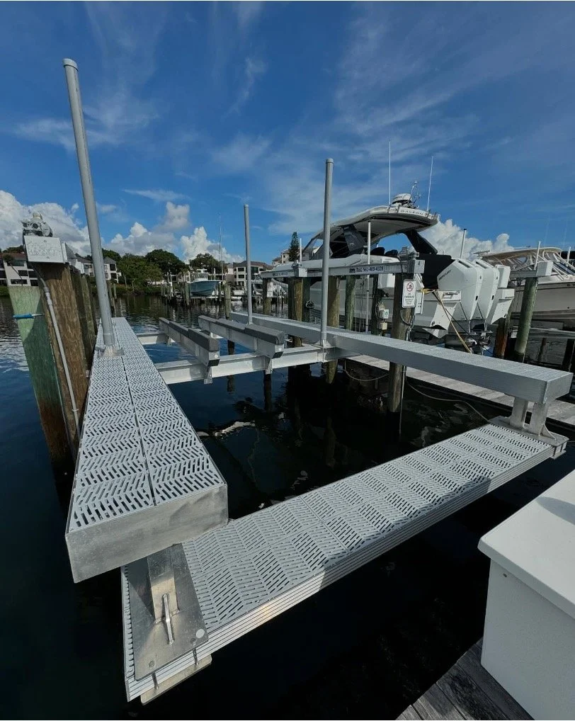 A boat dock with a metal gangway leading to a boat lift platform, with several boats docked on the water under a partly cloudy sky.
