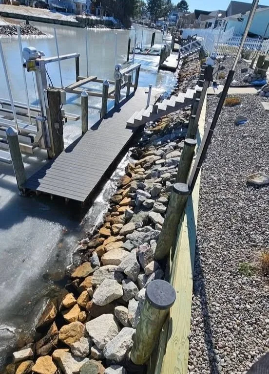 A dock with a ramp leading into a body of water, next to a rocky shoreline and a paved walkway with a wooden fence.