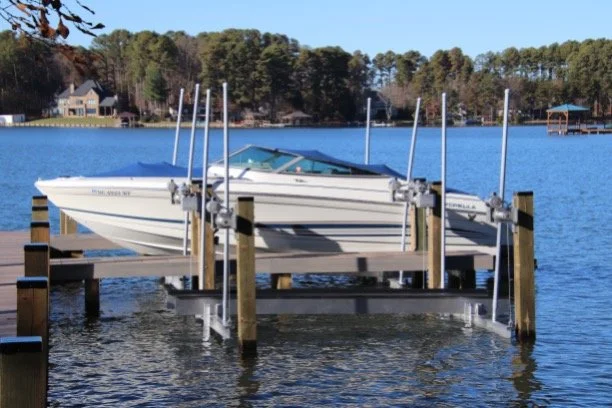A boat is on a boat lift at a lake, with houses and trees in the background.