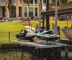 A dock with a jet ski and a boat covered with a tarp, located by a waterway with a grassy area and houses in the background.