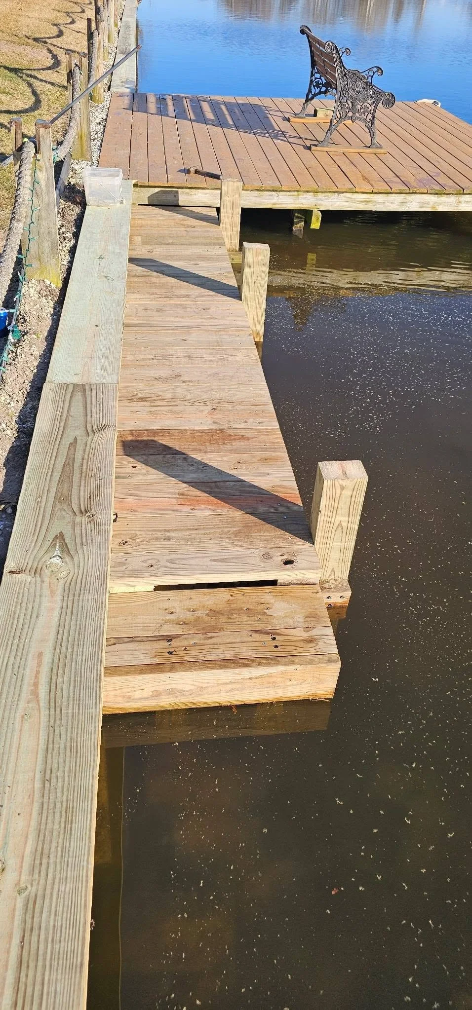 A wooden dock with a bench on it extending over a body of water. The area is sunny and clear, with reflections seen in the water.