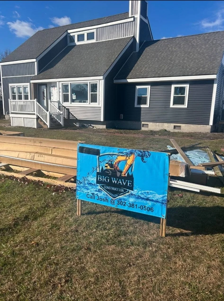 Construction site with a house in the background and a sign for Big Wave Construction in the foreground, along with building materials.