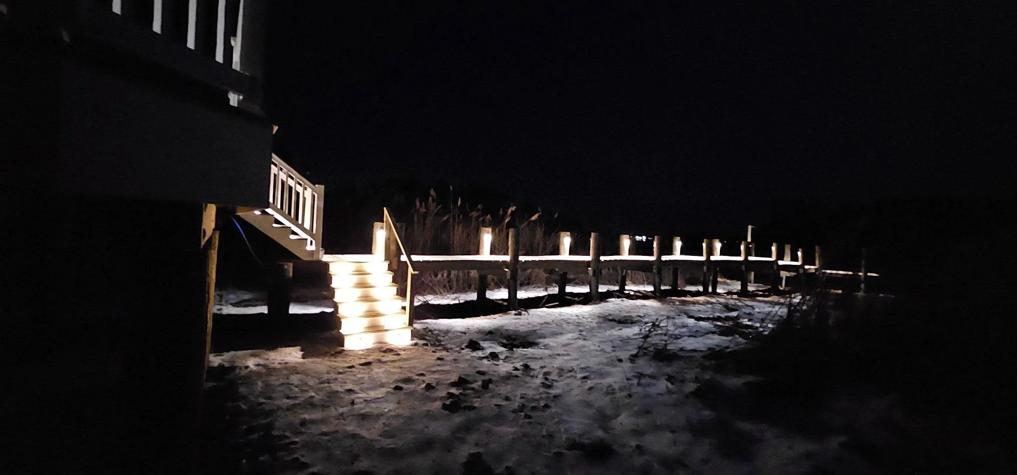 Nighttime scene of a wooden boardwalk with steps illuminated by small lights, leading to a sandy area and bushes, under a dark sky.