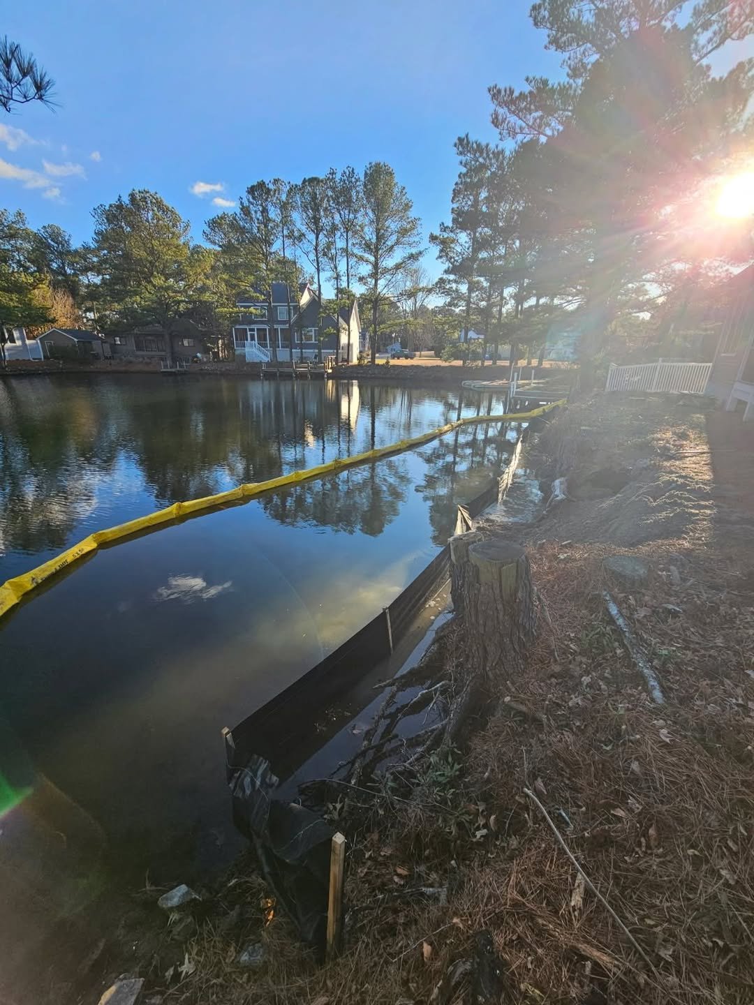 A small residential waterfront neighborhood with houses, trees, and calm water on a sunny day with clear blue sky.