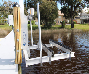 A boat lift with a metal frame and vertical support posts, installed in a lake or river, with trees and residential houses in the background.