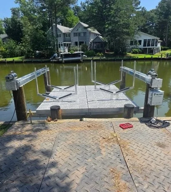 A small concrete boat dock with metal railings extends into a body of water, with a house and greenery in the background. The photo is taken during the day.
