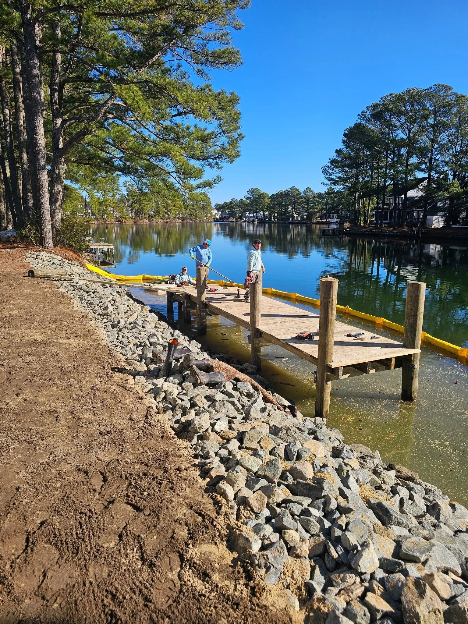 Workers installing a wooden dock along a lake shoreline with trees and houses in the background.