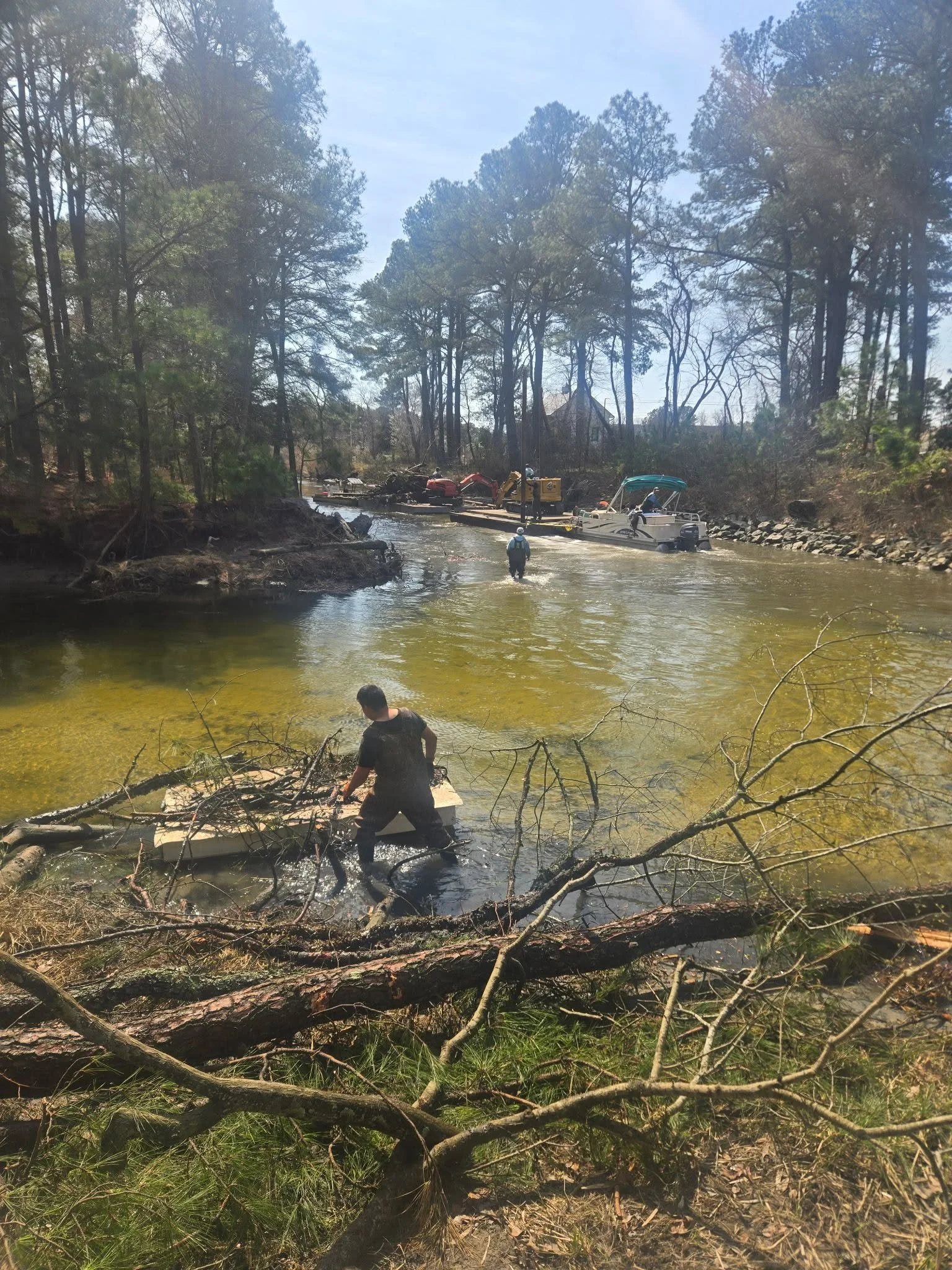 A scene of a flood in a wooded area with a person walking through shallow water, and a boat and construction equipment in the background.