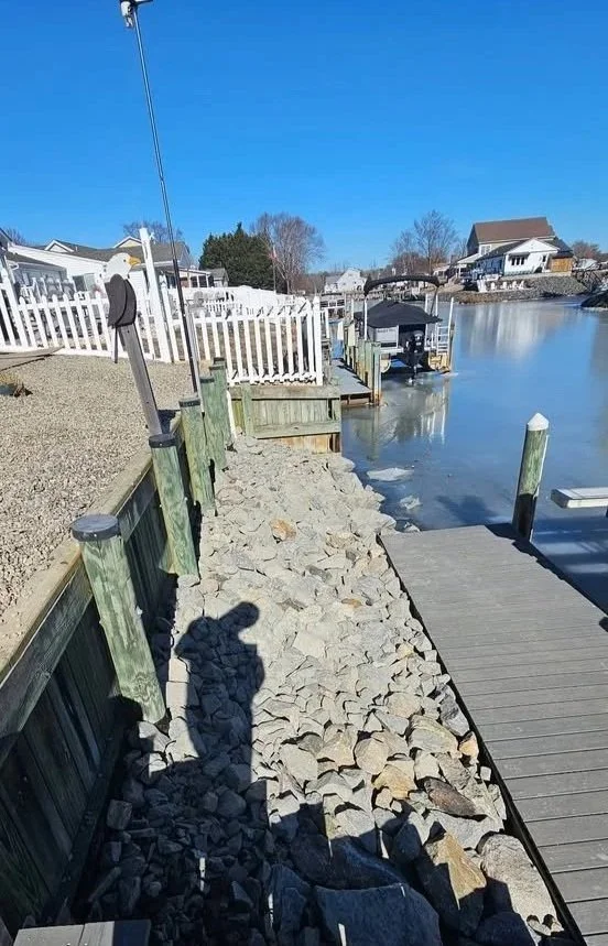 A rocky shoreline next to a dock on a calm waterway, with houses and trees in the background under a clear blue sky.