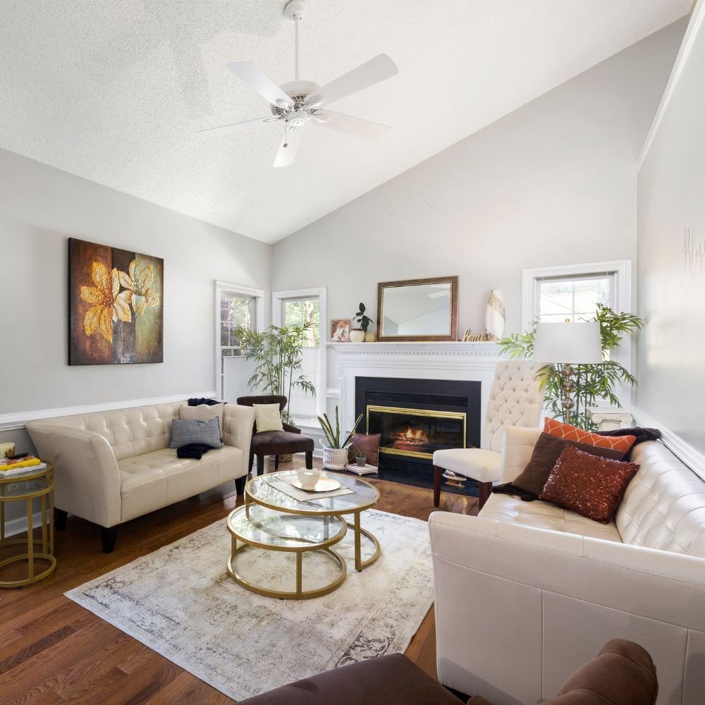 Living room with white sofas, a fireplace, a ceiling fan, a round glass coffee table, plants, and wall art.