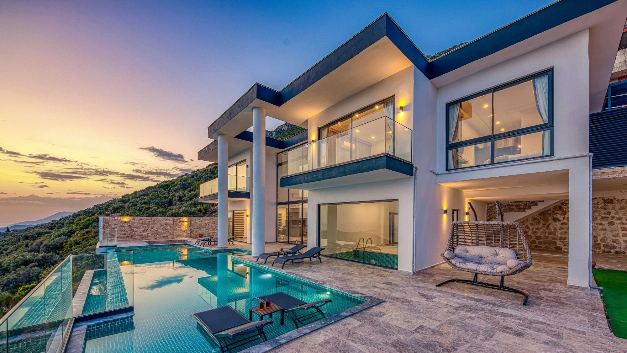 Luxury modern house with large glass windows, outdoor pool, and hillside view during sunset, with outdoor furniture and lighting.
