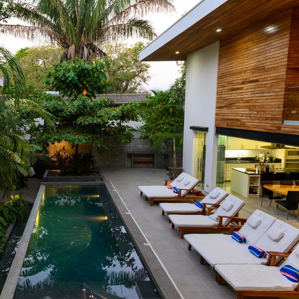 Modern backyard with a narrow swimming pool, four lounge chairs with white cushions and striped towels, and an open kitchen and dining area with wooden accents, surrounded by lush green trees.