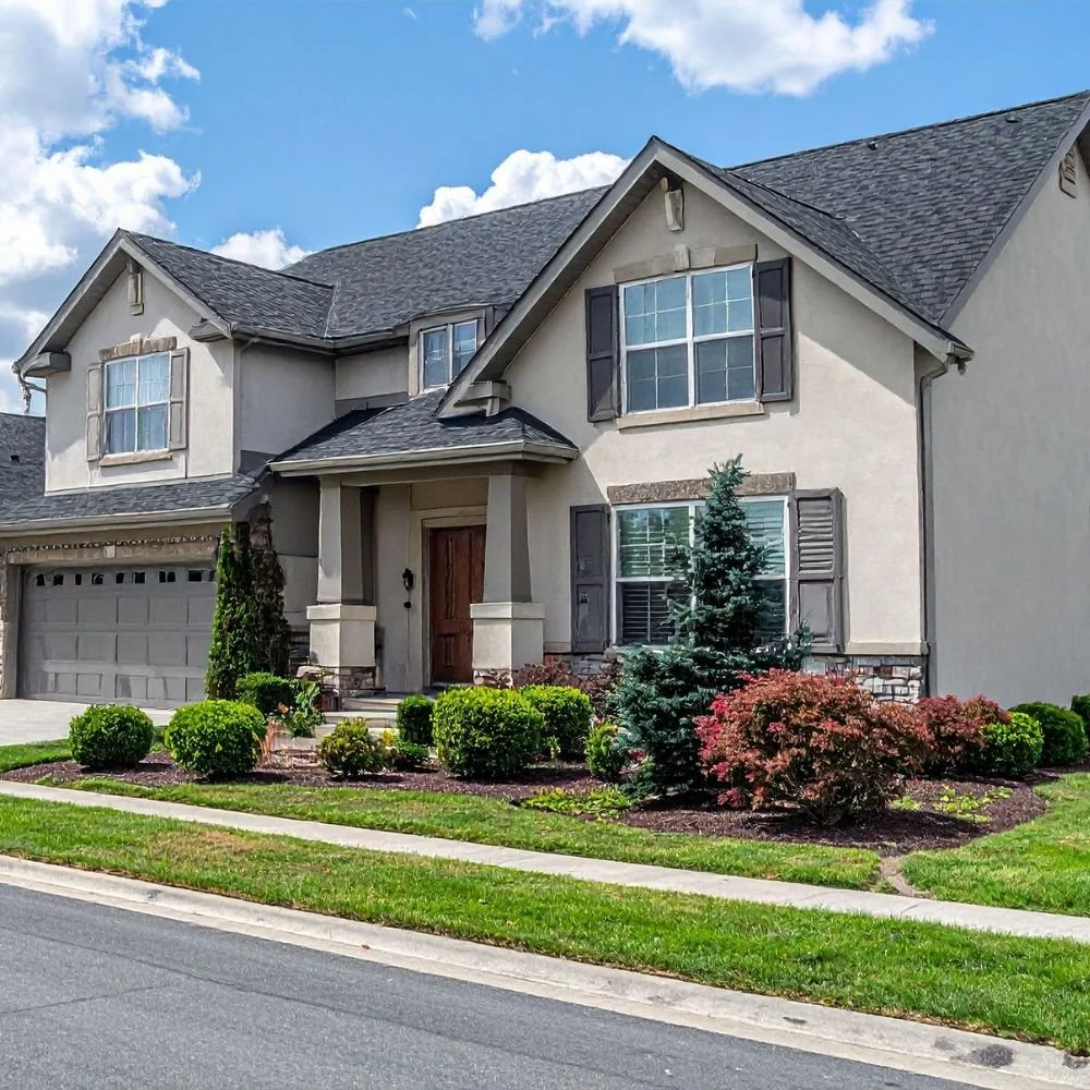 A two-story suburban house with a gray roof, beige exterior walls, and dark brown shutters. The front yard has neatly trimmed bushes, small trees, and a walkway leading to the front door. The sky above is partly cloudy.
