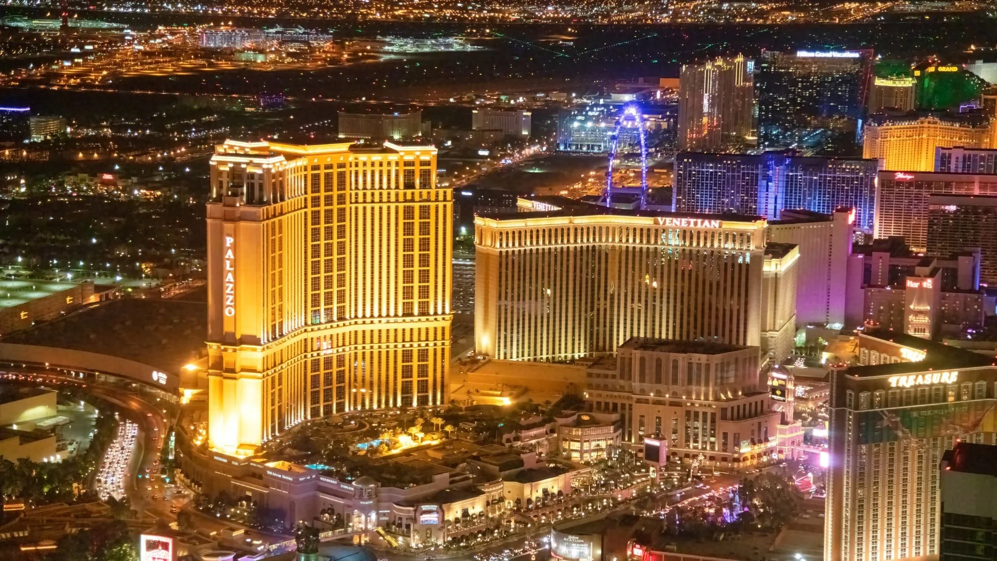 Nighttime aerial view of the Las Vegas Strip with illuminated hotels and casinos, including the Palazzo and Venetian, and a large Ferris wheel in the distance