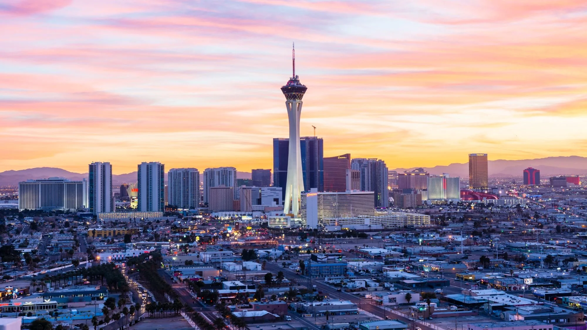Skyline of Las Vegas with the Stratosphere Tower during sunset, surrounded by city buildings and mountains in the background.