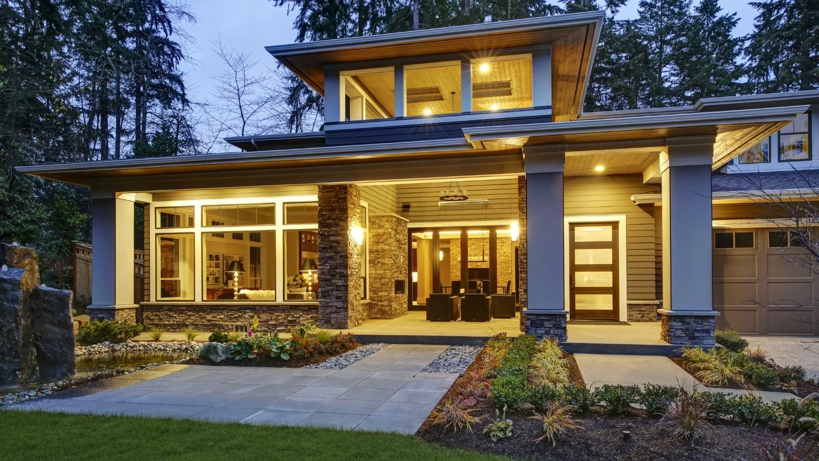 A modern, well-lit house with large windows, stone accents, and a front porch surrounded by landscaped garden and stone pathway, set against a wooded background during dusk.
