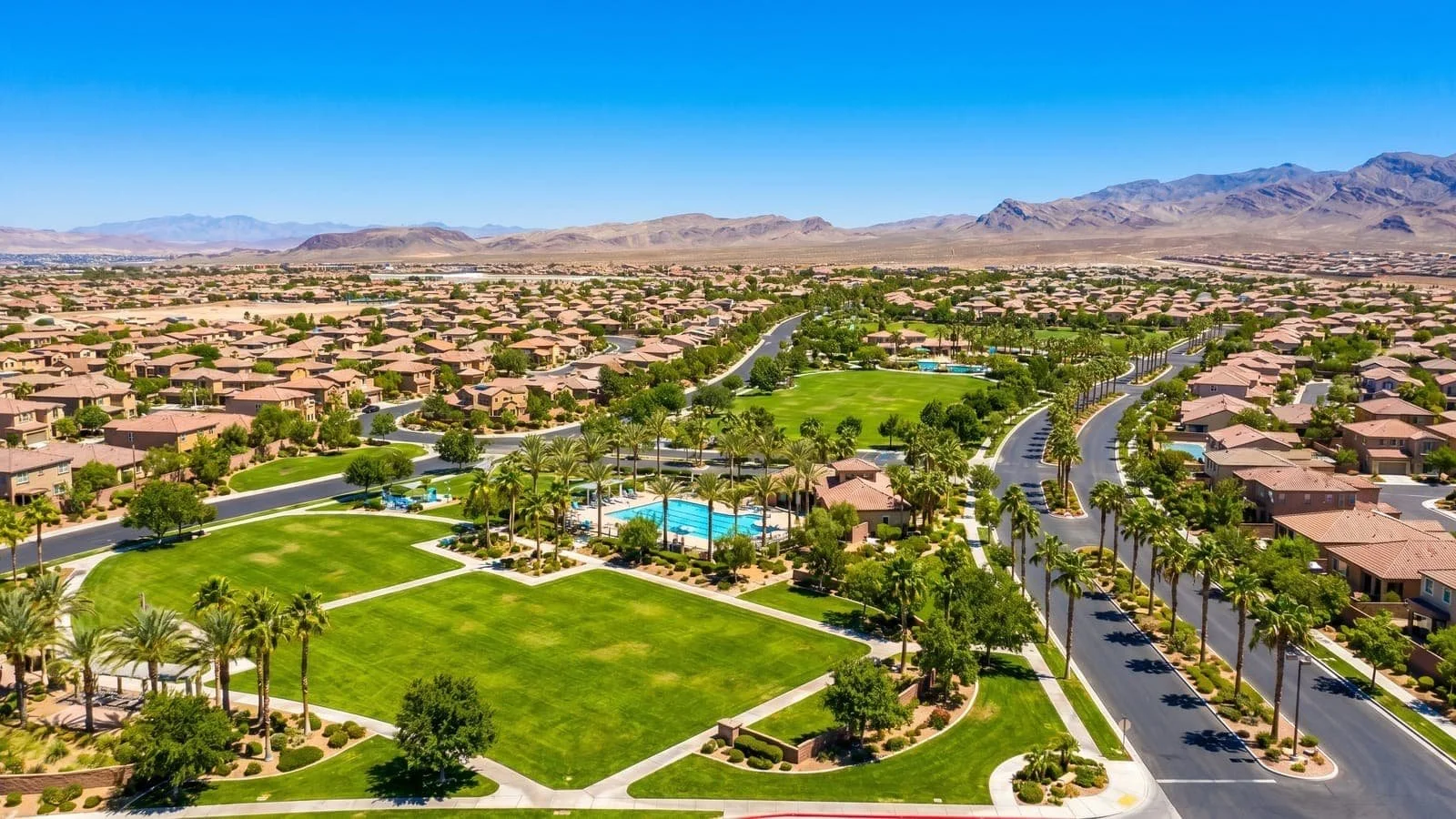 Aerial view of a suburban neighborhood with green parks, swimming pools, and tree-lined streets, surrounded by desert mountains under a clear blue sky.