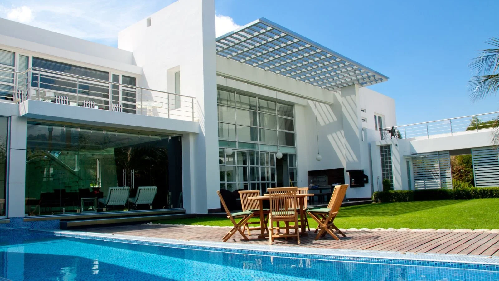 Modern white house with outdoor patio, swimming pool, wooden dining set, and lush green lawn under a sunny blue sky.