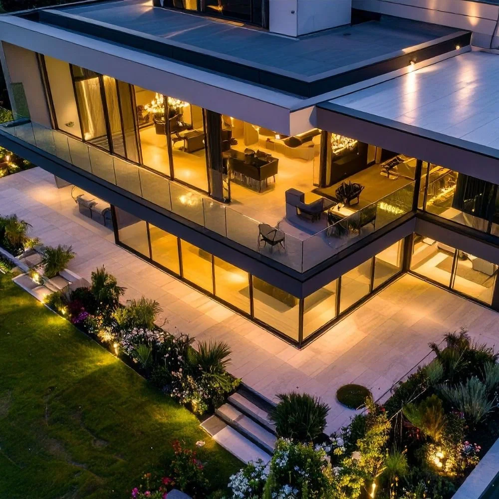 An aerial view of a modern multi-story house with large glass windows, illuminated from within, surrounded by a landscaped garden with pathway lights, plants, and trees.