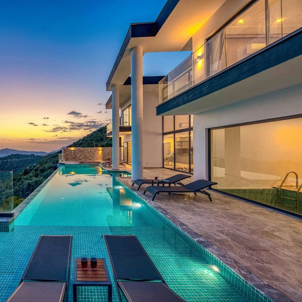 Modern house with infinity pool on hillside at sunset, glass balcony, and mountain view, with lounge chairs on deck.