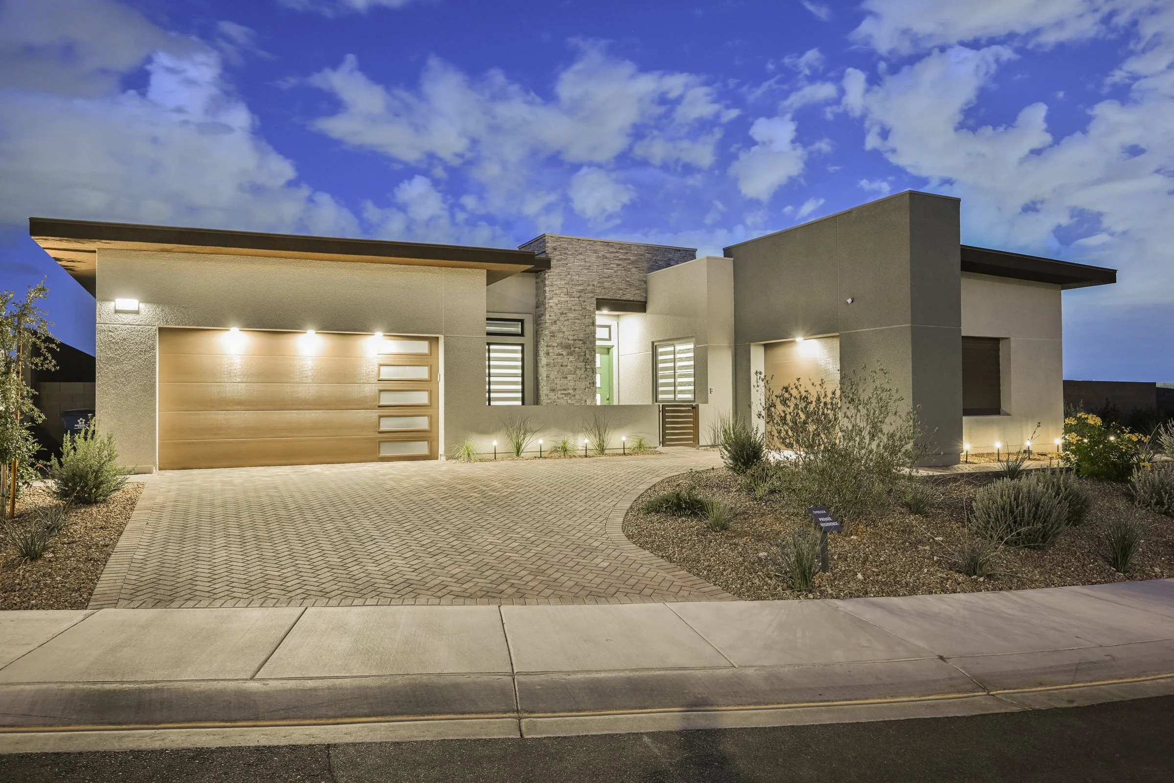 Modern house with a flat roof, stone and stucco exterior, illuminated by outdoor lighting, with a paved driveway and desert landscaping, under a partly cloudy evening sky.