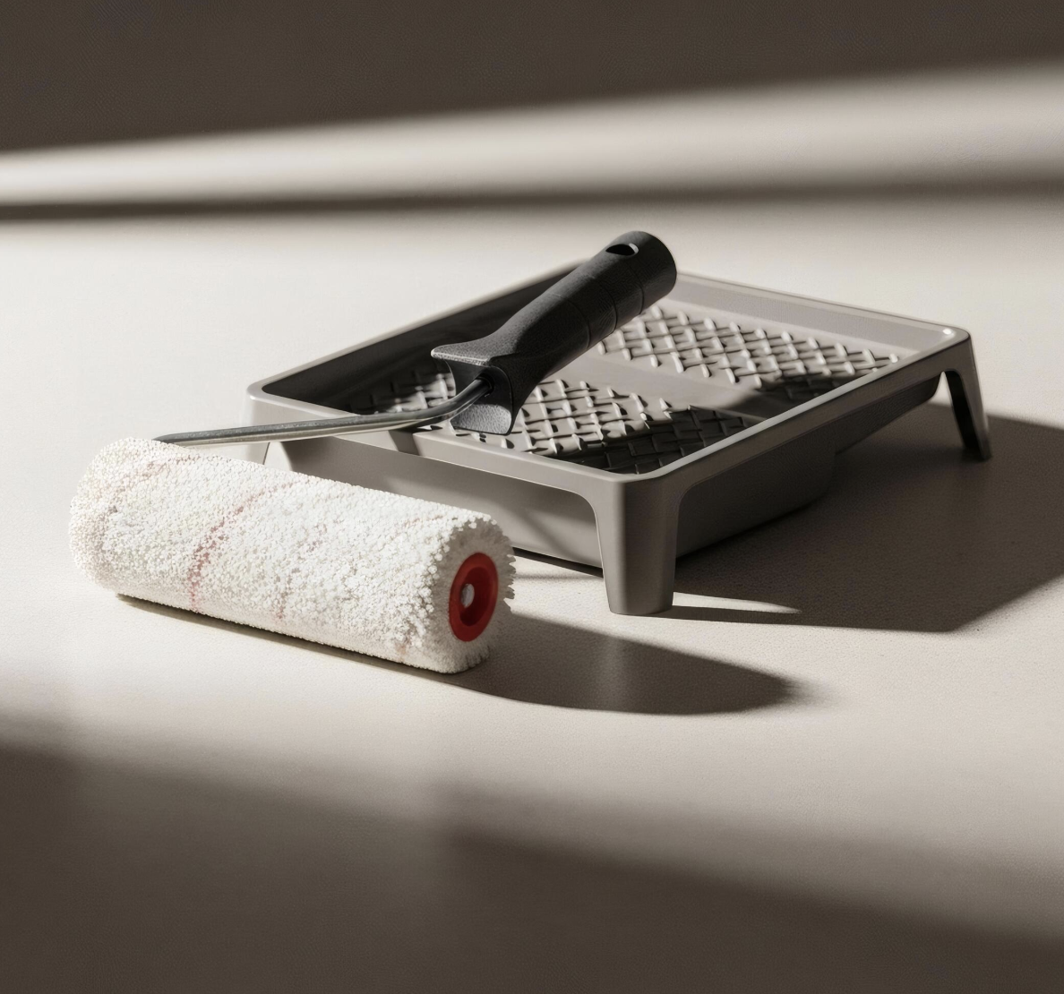 A paint roller, a grout float, and a paint tray with grid pattern are on a flat surface with shadows and light