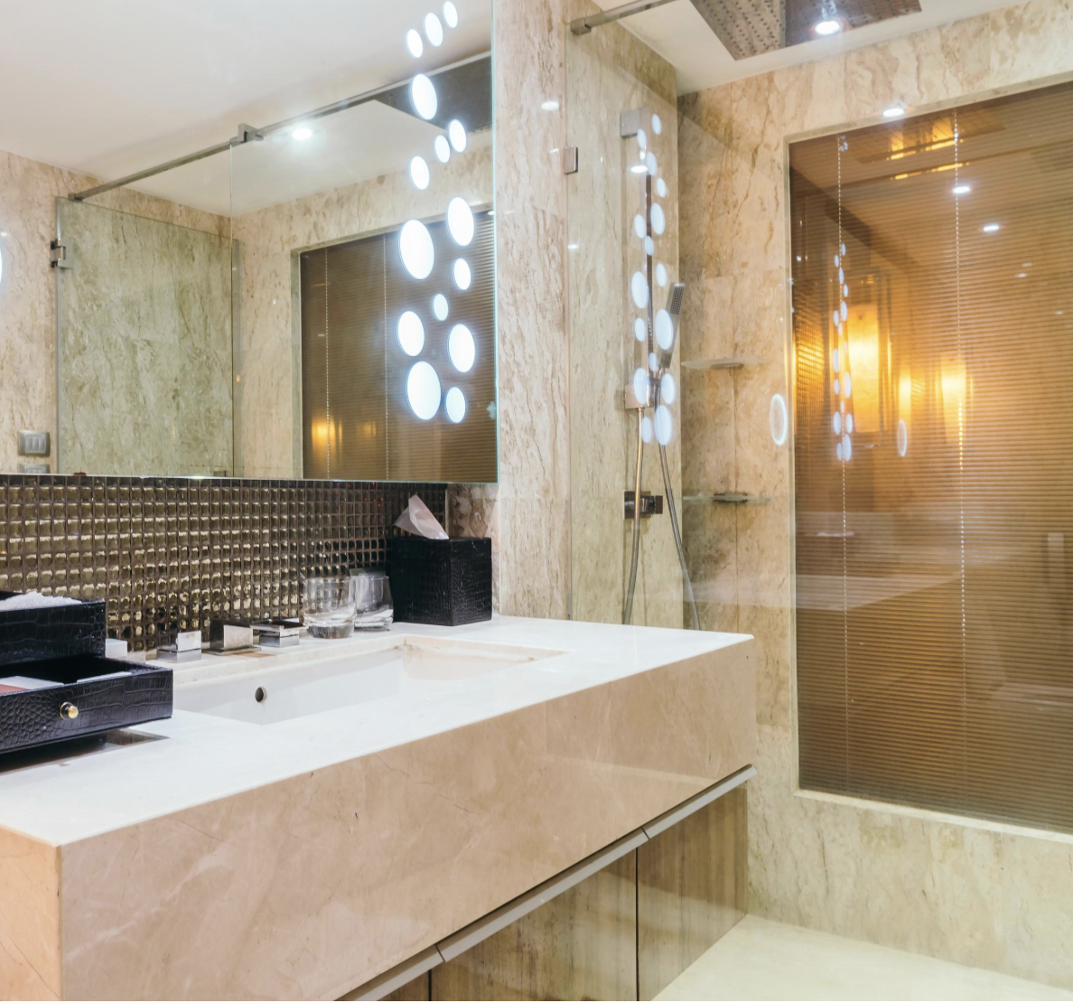 Modern bathroom with marble walls and countertops, large mirror, decorative lighting, and a glass shower enclosure with a showerhead and shelves.