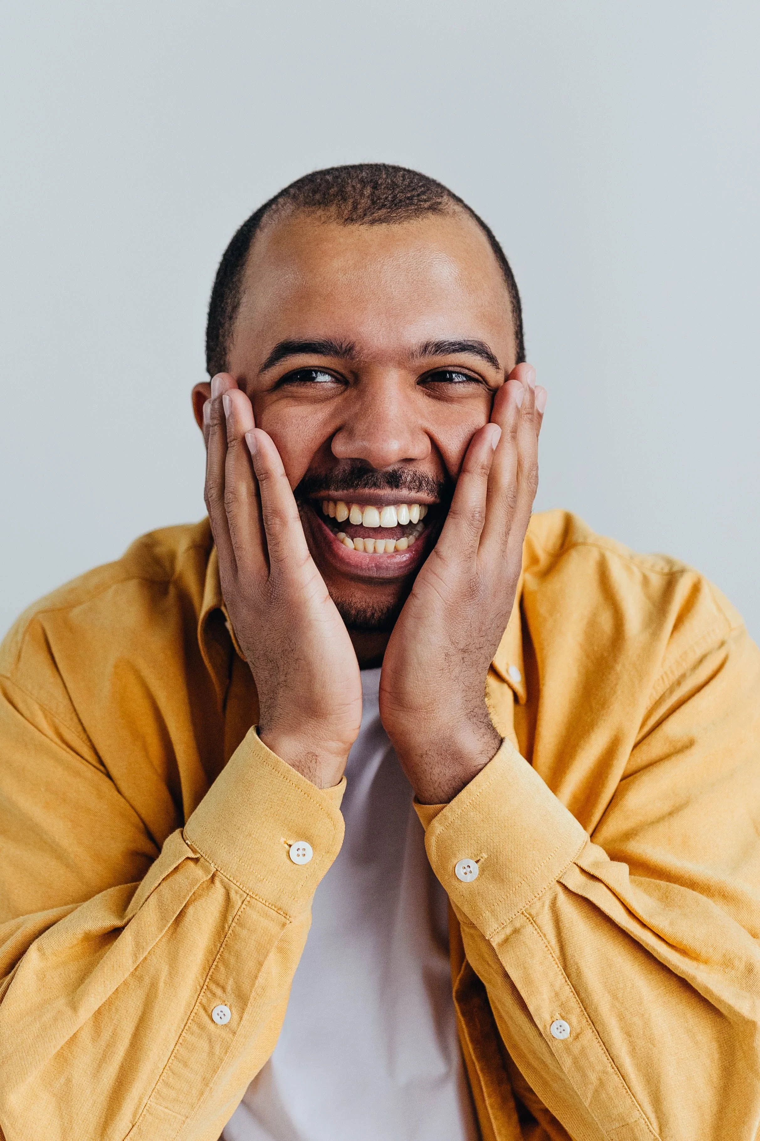 A man with a shaved head, beard, and a big smile, holding his face with both hands, wearing a yellow shirt and a white T-shirt underneath, against a light gray background.