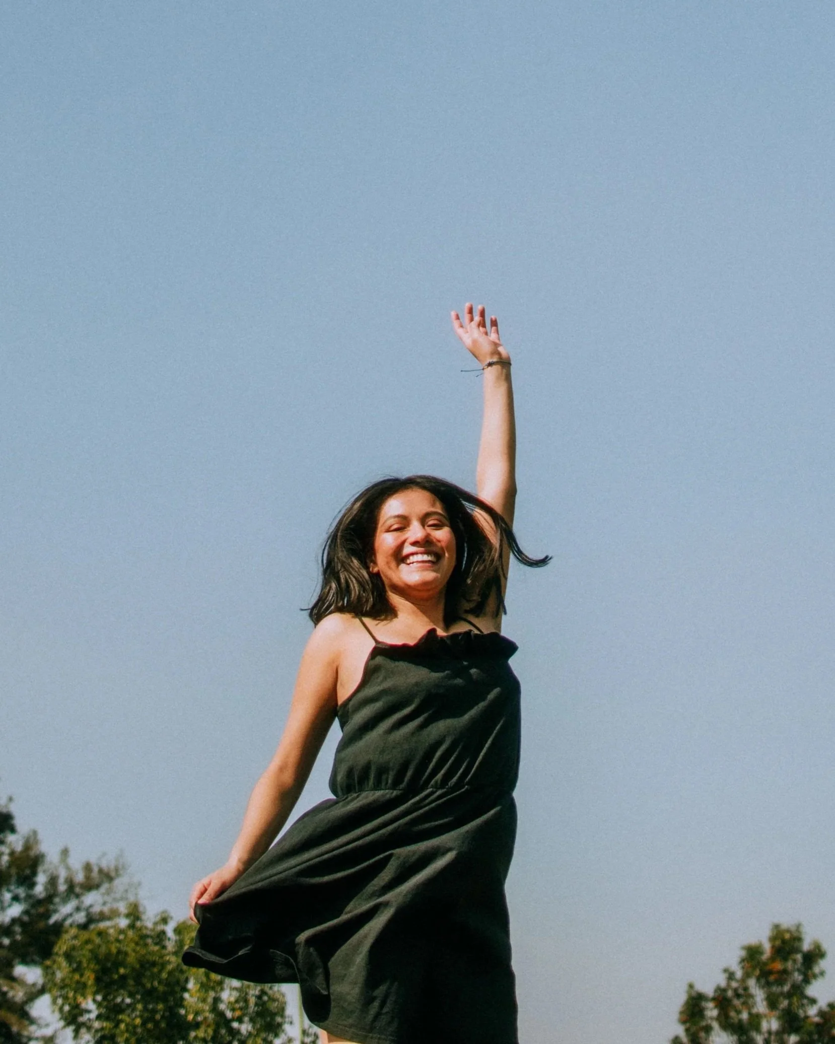A woman with long brown hair smiling and wearing a black dress with thin straps, standing outdoors with trees and a clear blue sky in the background, raising one arm in the air.