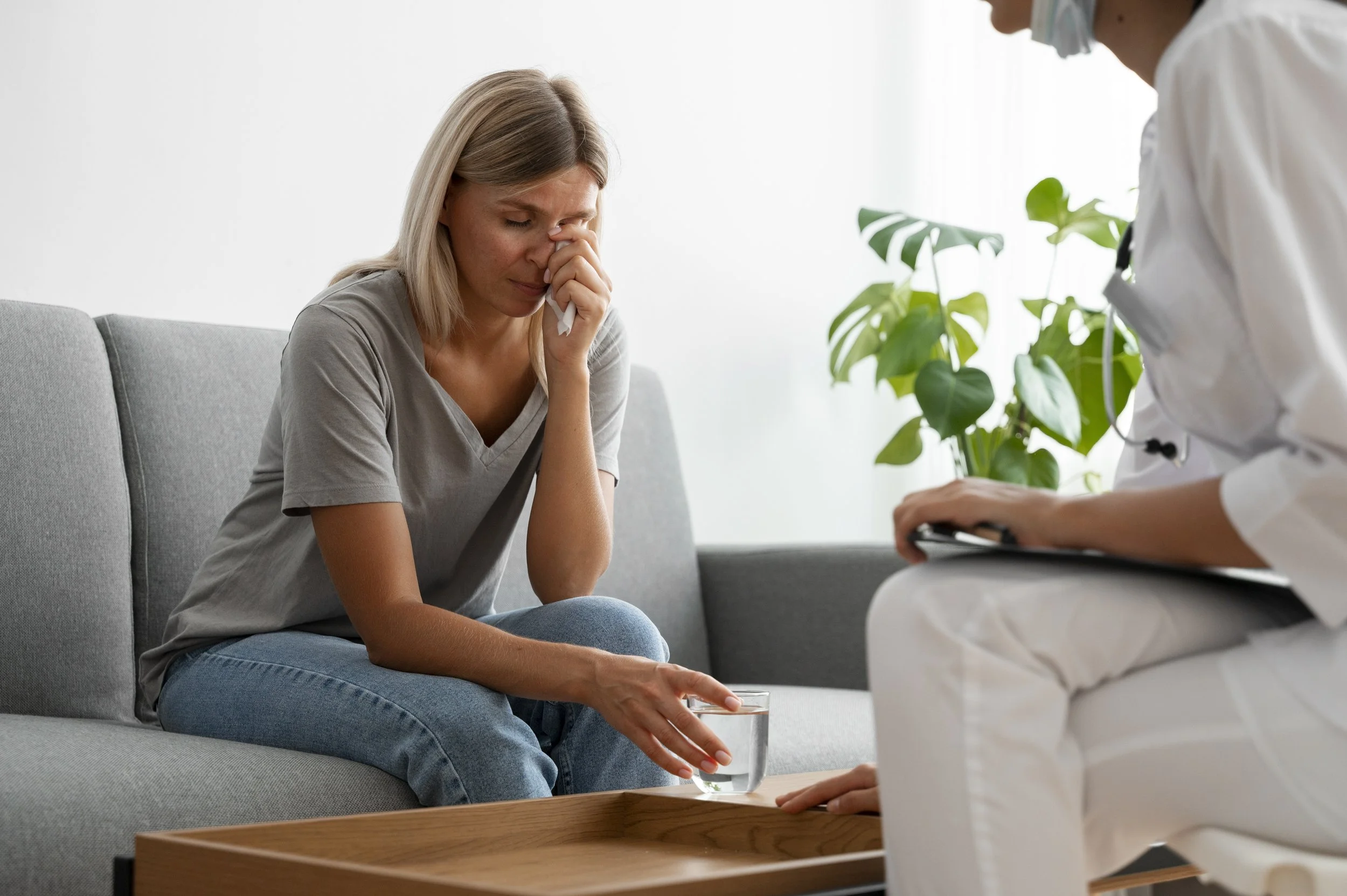 A woman sitting on a gray couch, appearing distressed, with a tissue in her nose, holding a glass of water on the table in front of her. A healthcare professional wearing a white coat and face mask is attending to her.