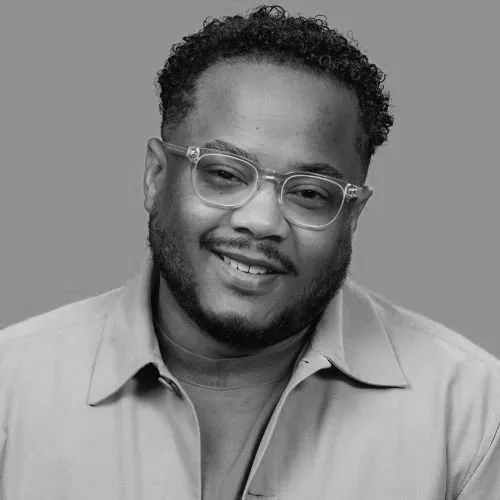 A portrait of a young man with curly hair, beard, wearing glasses, and smiling, dressed in a button-up shirt against a plain background.