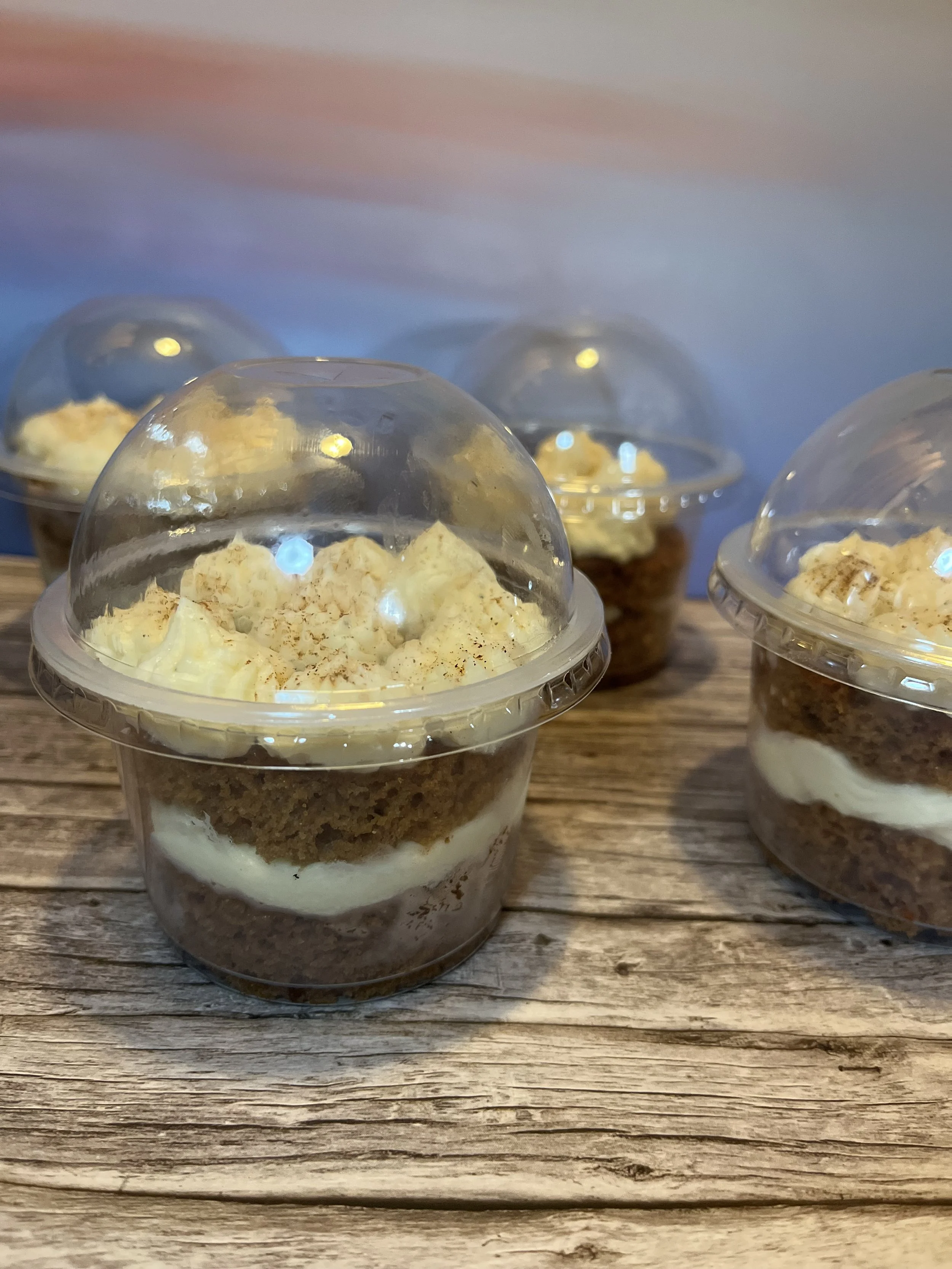 Four cups of layered dessert with crumbly graham cracker crust, creamy filling, and topped with whipped cream and sprinkled spices, placed on a rustic wooden surface.