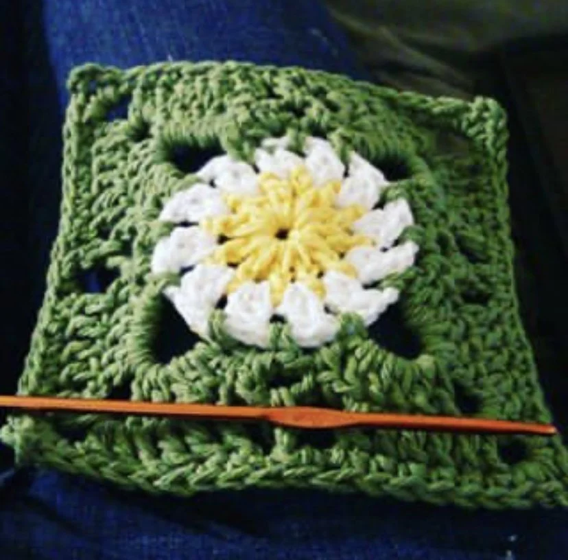 A green crocheted square with a white and yellow flower pattern in the center.