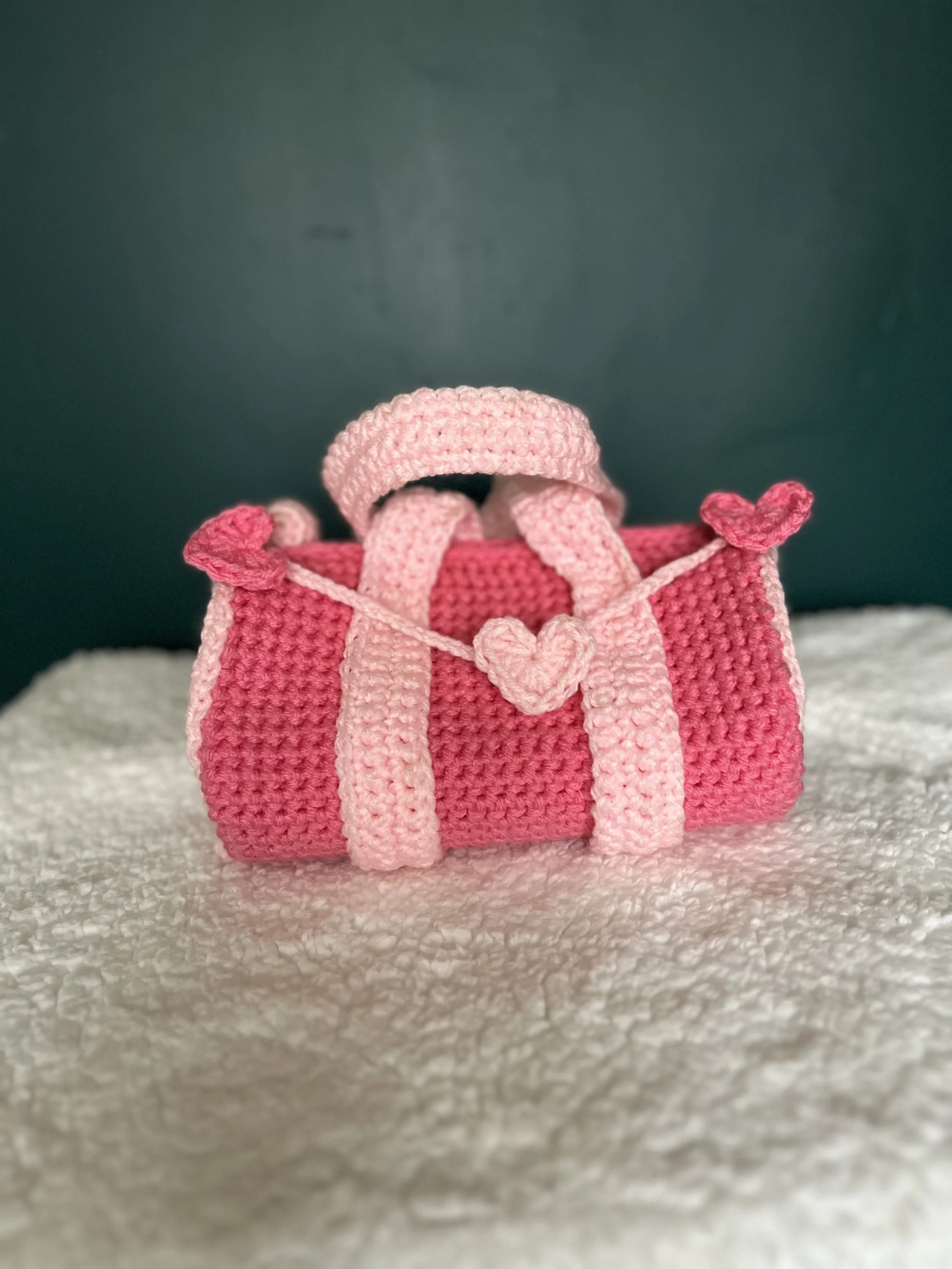 Handmade pink and light pink crocheted gift bag with a heart-shaped decoration, placed on a soft white surface with a dark green background.