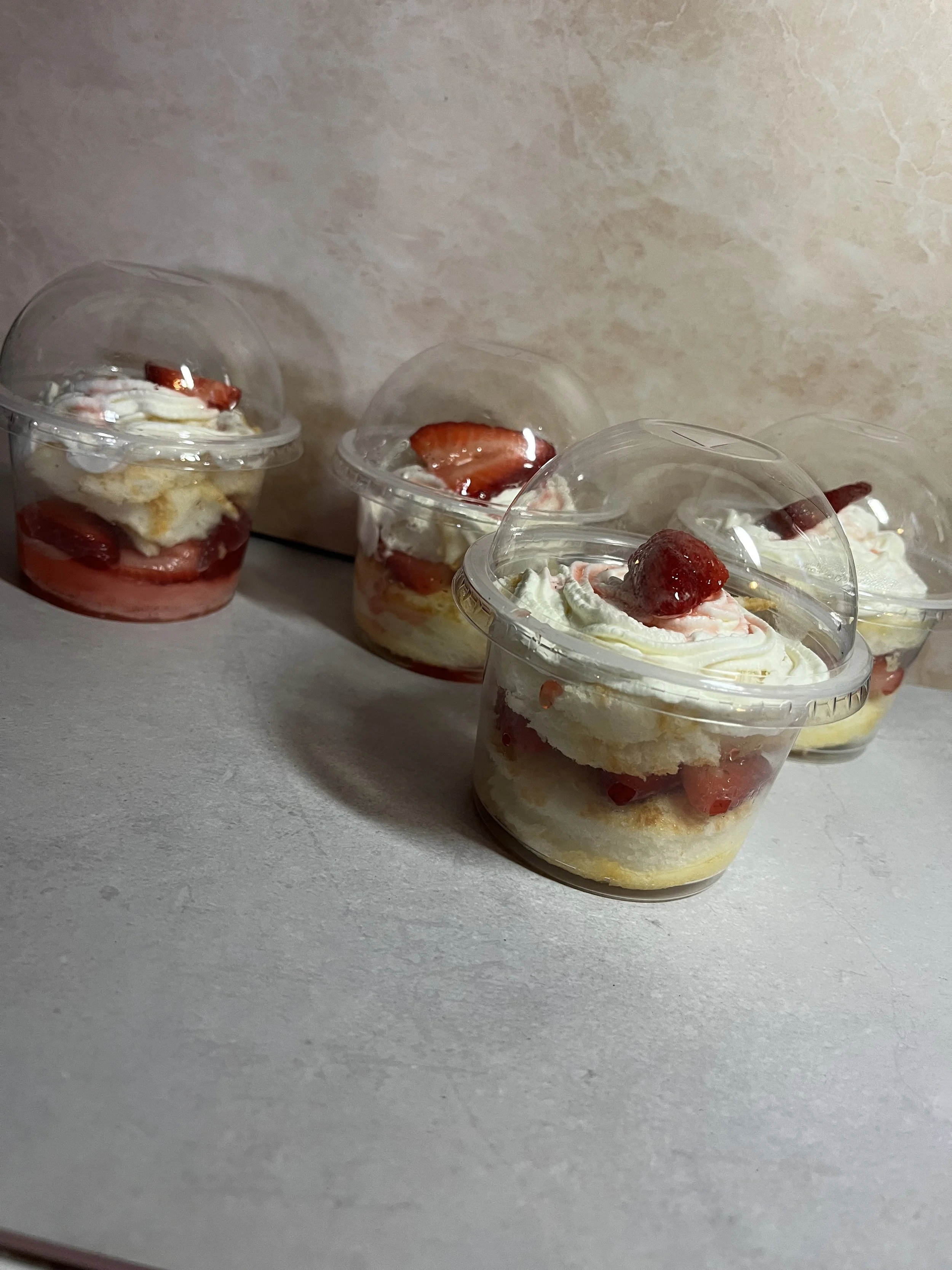 Clear plastic cups filled with layered strawberry shortcake, topped with whipped cream and a fresh strawberry, on a white surface.