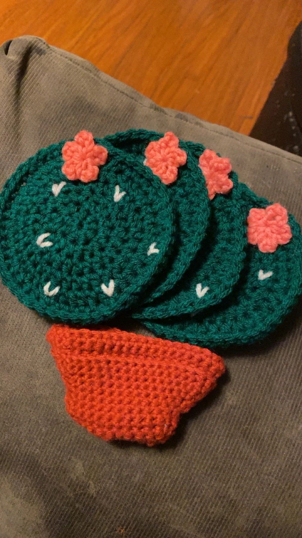 Set of four crocheted coasters in green with pink flowers and a small orange wrapped crochet item, placed on a gray fabric surface.