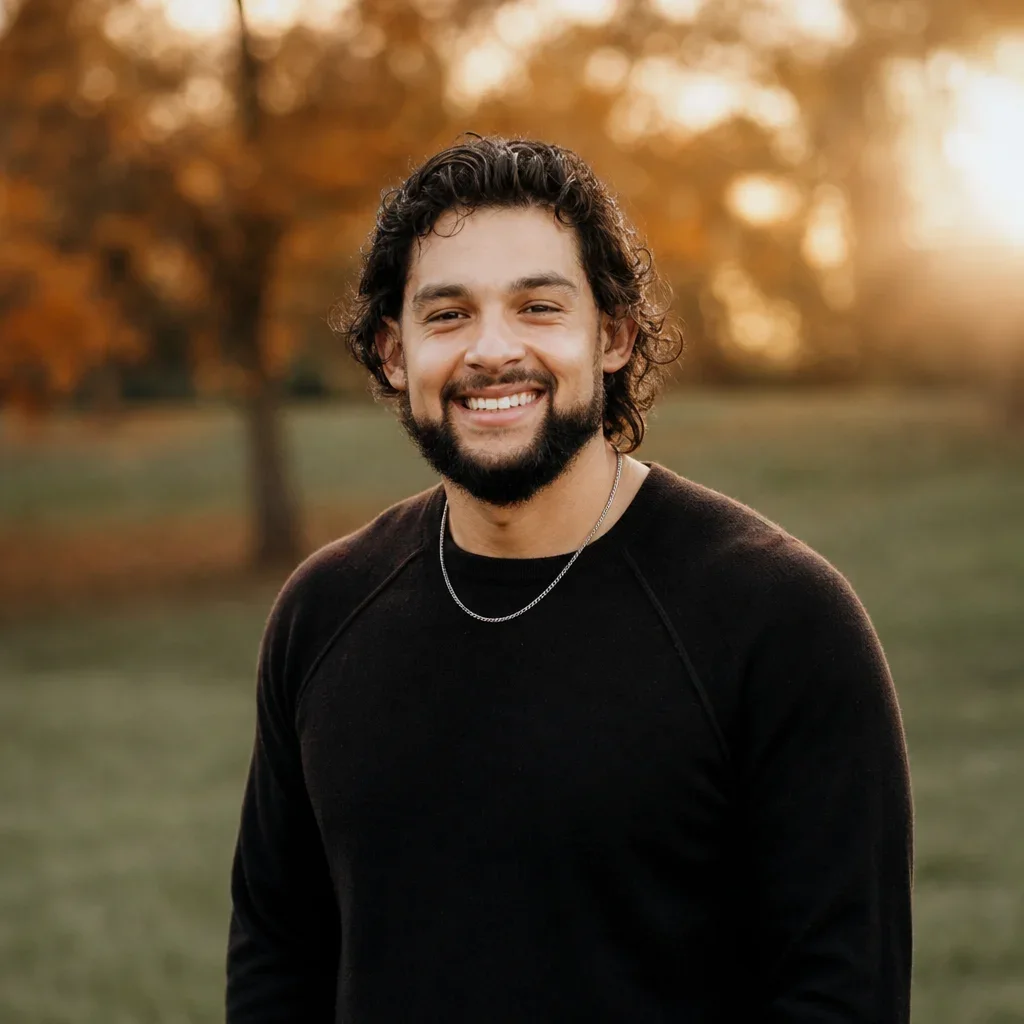 A smiling man with curly hair and a beard standing outdoors during sunset, wearing a black long-sleeve shirt and a silver chain necklace.